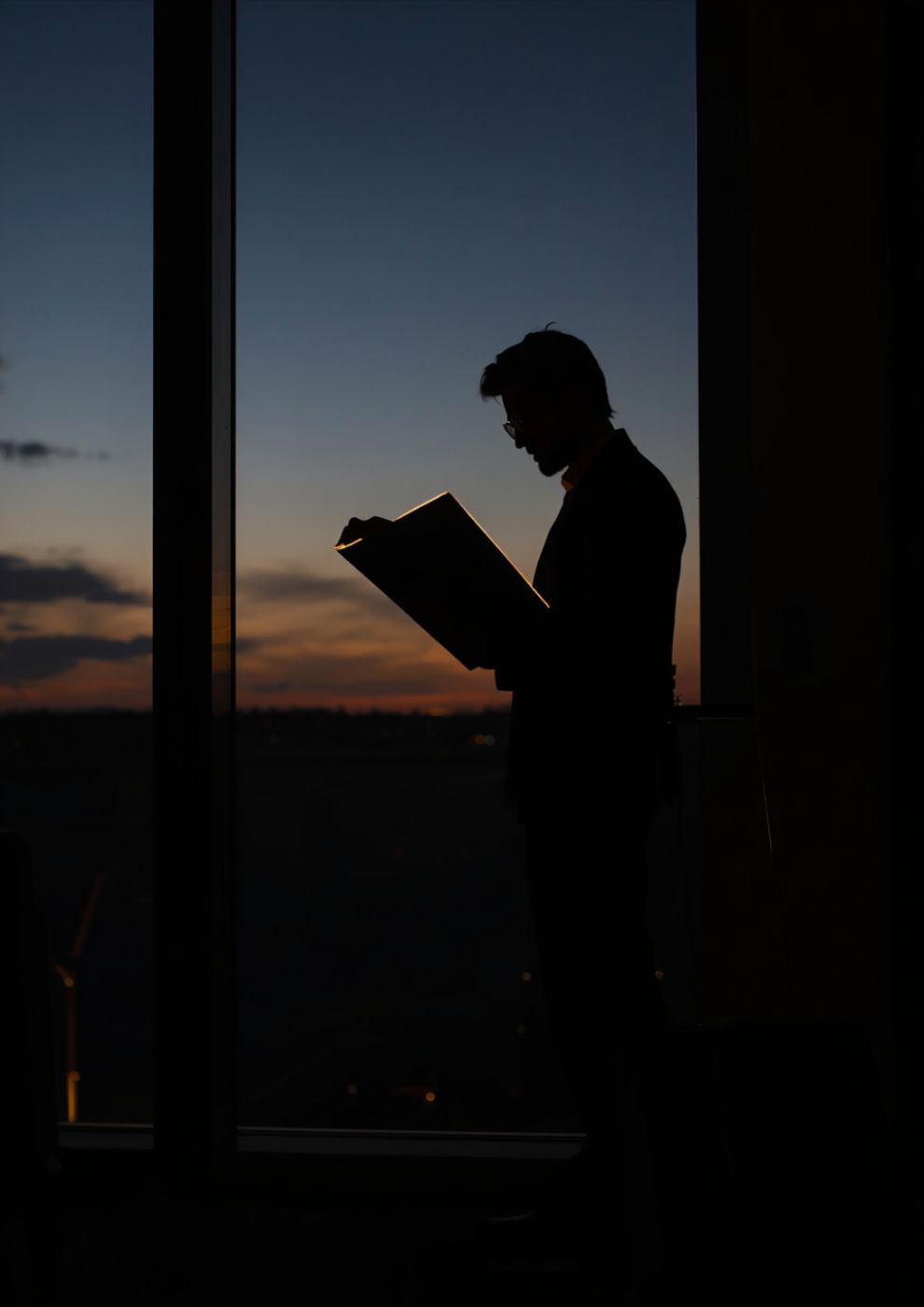 A silhouette reading a document by a window at sunset.